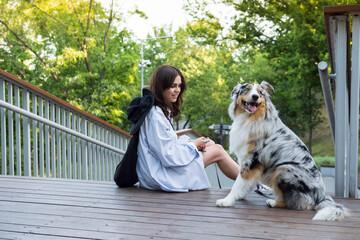 Young woman sitting next to her cute aussie dog in a park. Blue merle australian shepherd dog in urban park area next to female owner, walking pets in the city