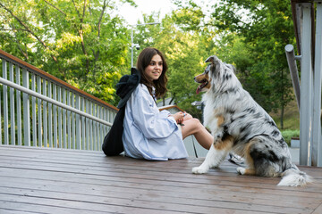 Young woman sitting next to her cute aussie dog in a park. Blue merle australian shepherd dog in urban park area next to female owner, walking pets in the city