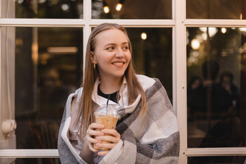 Happy smiling blonde woman sitting in cafe, enjoy of ice latte in to go cup, covering her shoulder plaid. Girl look dreaming and thoughtful, enjoy of coffee break after evening after work.