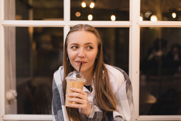 Happy smiling blonde woman sitting in cafe, enjoy of ice latte in to go cup, covering her shoulder plaid. Girl look dreaming and thoughtful, enjoy of coffee break after evening after work.