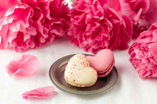 Pink coral peony flowers and french sweet cookies macarons macaroons on linen table cloth background. Heart shape macarons and bouquet of peony.
