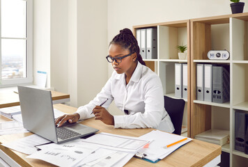 Businesswoman working in her office. Young African American woman in a white shirt and glasses sitting at her desk, using a modern laptop computer, doing paperwork, working with business documents