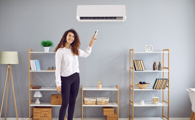 Smiling young woman holding remote controller switching on air conditioner. Cheerful businesswoman operating air conditioner setting comfortable temperature in office or home
