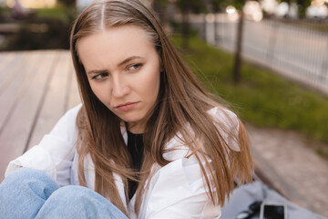 Sad tired young lady in shirt and jeans sits on bench, thinks, suffer from headache and migraine in...