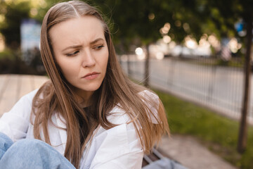 Sad tired young lady in shirt and jeans sits on bench, thinks, suffer from headache and migraine in park. Emotions, health care problems, stress and pressure, insult, offense girl hugs her legs.