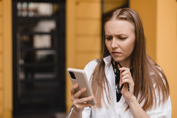 Confused puzzled blonde woman in casual clothes, standing near yellow building, holds a smartphone...