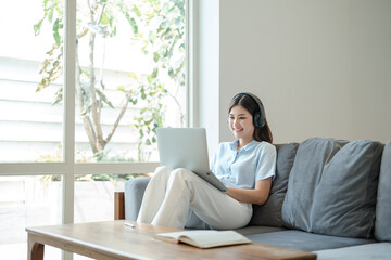 Beautiful Young asian woman at home sitting on the sofa using laptop and headphone listening to music at home.