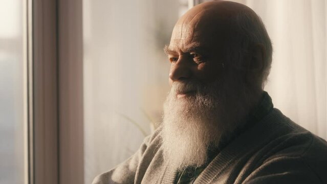 Happy senior man with a beard standing near window, old age, good memories