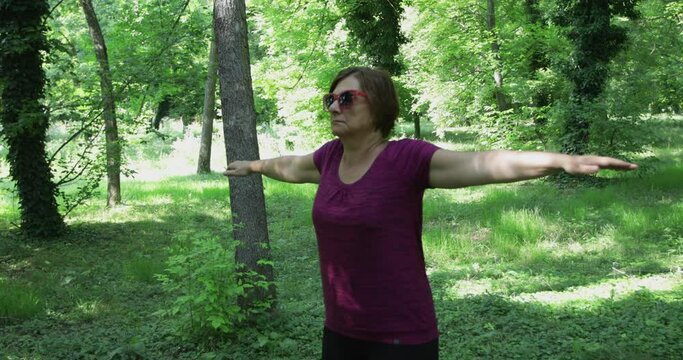 50 year old mid adult woman doing neck and upper spine exercise outdoors, making circles shoulder joints forward and backwards with arms stretched, real people, steadicam