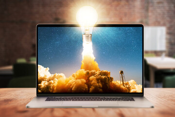 Concept light bulb glows and successfully lift off with smoke and clouds from the laptop display on wooden desktop in office. Successful start up, creative thinking and think differently