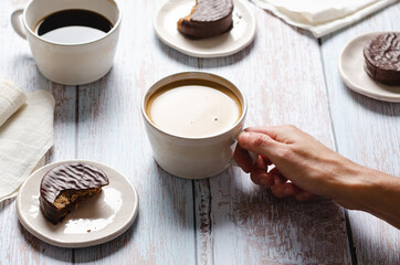 Girl drinking coffee and eating chocolate alfajores on a wooden table.