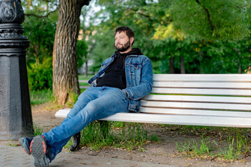 Obraz premium attractive caucasuan bearded young man sitting on bench in park looking aside