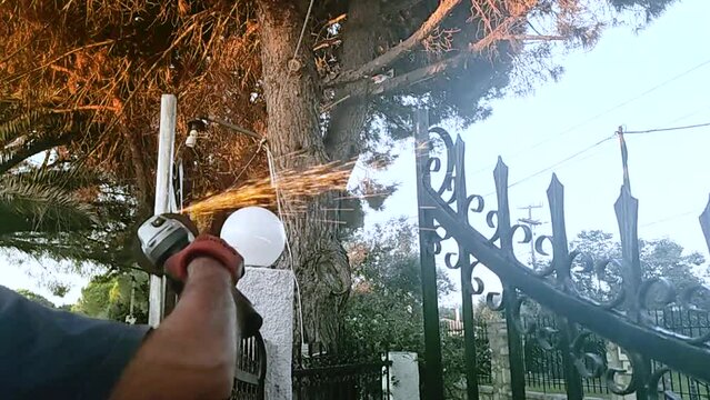 Caucasian Man is Busy Using Electric Grinder Tool to Smooth an Iron Gate. Fixing the Entrance Door of a House with Pine Trees. 4K Video Footage