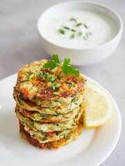 Pile of vegetarian fritters or vegetable cutlets from zucchini (courgette) with fresh herbs on top. Lemon and bowl of yogurt sauce as garnish.