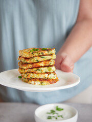 vegetable pancakes or fritters piled on a plate in a woman's hand, out of focus a bowl with yogurt...