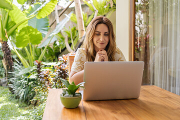 Young beautiful woman sitting in a garden with laptop in a tropical resort. Girl with laptop computer connecting to 4G wireless for browsing social media working remotely at terrace