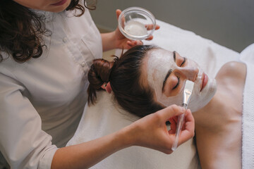 Obraz na płótnie Canvas Cosmetologist applying facial cream all over young woman's forehead at spa salon. Skin treatment. Girl facial treatment.