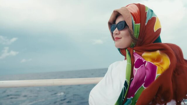 Woman on a sea voyage on a windy day. Korean female person sitting on upper deck of cruise yacht enjoying wind and smiling. Girl wearing bright shawl on her hair and sunglasses. Summer vacation moment