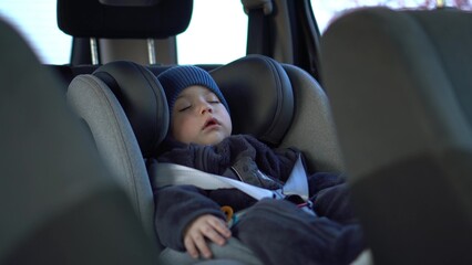 A small child sleeps in a car seat in a parking lot. Boy in a sweatshirt and hat.