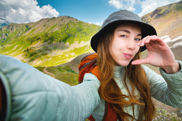 Attractive young caucasian woman in panama, down jacket and backpack taking selfie and half heart gesture high in mountains looking and winking at camera.