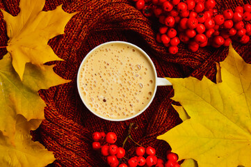 A cappuccino cup on the background of a brown knitted sweater. Yellow maple leaves and red rowan berries. Warm and cozy autumn. Leaf fall.