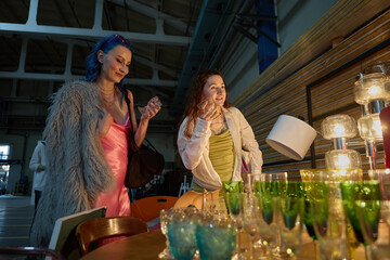 Two young diverse women in fashionable clothes buying vintage wine glasses in a thrift shop. Portrait of a LGBT couple shopping on a flea market