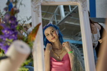 Beautiful queer person with dyed blue hair trying a vintage necklace in a thrift shop. Portrait of a cheerful LGBT female picking fashion accessories on a flea market sale
