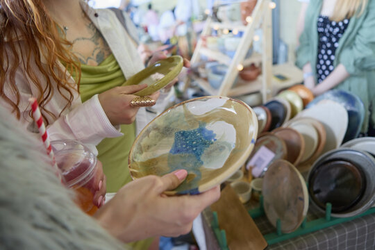 Two diverse females in fashionable dresses buying handmade pottery and dishware at the flea market sale. Young LGBT couple shopping for vintage and hand crafted plates in a store