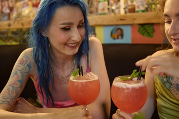 Cheerful blue haired female drinking cocktails with her girlfriend in a bar. Portrait of a happy LGBT couple of tattooed white women spending time together