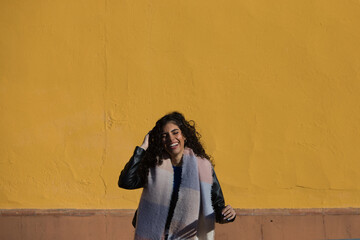 Obraz na płótnie Canvas pretty young woman with curly brunette hair against a yellow background is dressed in winter clothes and wearing a scarf to protect herself from the cold. The woman is happy and having fun.