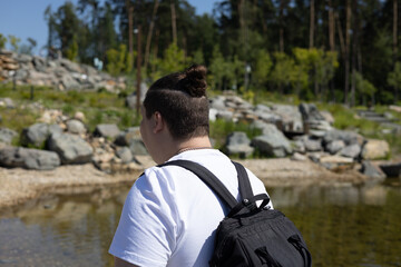 Fototapeta premium A male traveler with a backpack stands with his back to the camera against the backdrop of a mountain pond strewn with stones. In summer, a man travels through a mountain park.