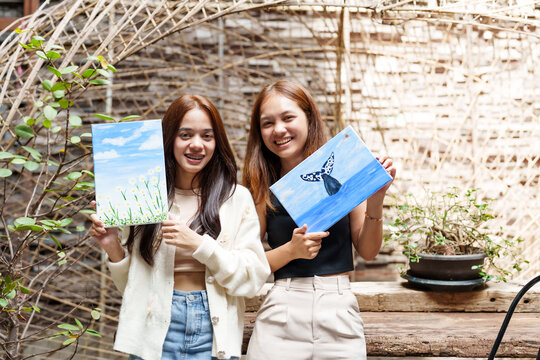 Close up. Two Asian female apprentice painters. Hold painting in hand, brush painting as recommended by art teacher, whale, sea and field of flowers in the school art classroom. - Powered by Adobe