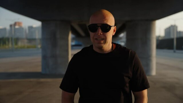 Happy brutal man in black glasses walking through the car park under the bridge. A sunny day in the city.