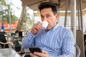 Mature latin man drinking coffee with cell phone in hand at street cafe