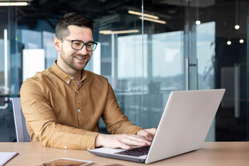 A smiling young businessman works in the office at a laptop. He sits at the table and types information, chats with clients, partners, communicates online