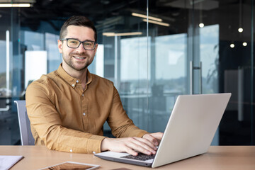 Portrait of a young male student confidently sitting at a desk in the office and smiling at the...
