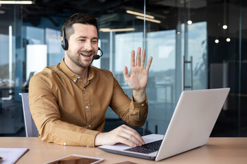 A young businessman in a headset smilingly talks on a video call on a laptop. Sitting in the office at the table and holding a business meeting, training, greeting and waving at the camera - Powered by Adobe