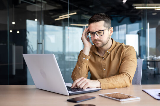Problems at work. Pensive and serious male office worker, sitting at a desk in front of a laptop, holding his head with his hand and looking worriedly at the screen