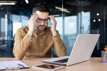 A worried young male businessman sits at a desk in the office in front of a laptop and holds his head in his hands. Thinks about solving financial problems and mistakes, upset by the results - obrazy, fototapety, plakaty