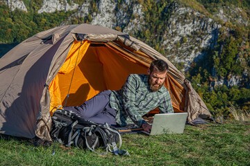 The concept of remote work or freelancer lifestyle. Young man using computer notebook to check news in front of a tent while camping on a holiday