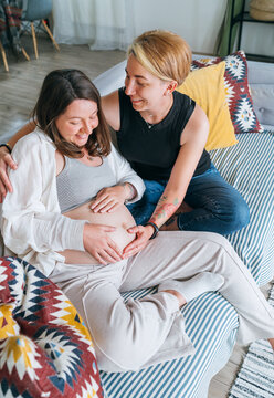 Young woman tender touching partner's female pregnant belly. Same-sex marriage couple in the home living room.  Woman's health, happy pregnancy doula supporting and calm mental mood concept image.