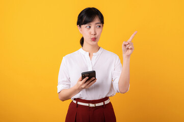 Portrait beautiful young asian woman enterpriser happy smile wearing white shirt and red plants pointing finger gesture to free copy space and using smartphone isolation on yellow background.