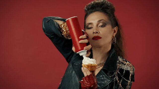 A woman applies whipped cream from an aerosol can to a cupcake, then squeezes the whipped cream into her mouth, chews and winks. Portrait of a woman on a red background close up. Advertising.