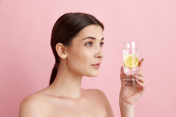 Beautiful young woman with healthy, well-kept skin taking care after health, drinking water with lemons against pink studio background. Concept of natural female beauty, body and skincare, health, ad