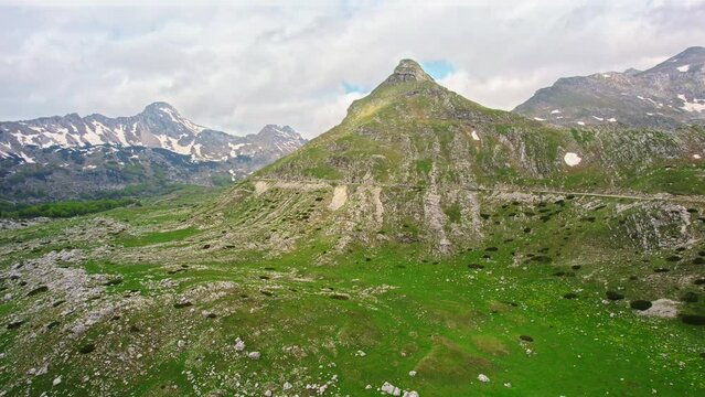 A mountain peak around which the road twists, shot from a drone in spring, behind which the highest mountain peaks with remnants of snow can be seen.