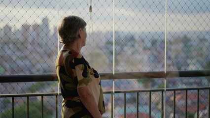 Older retired woman standing at apartment balcony looking at city view. Senior female person in 70s overlooking cityscape