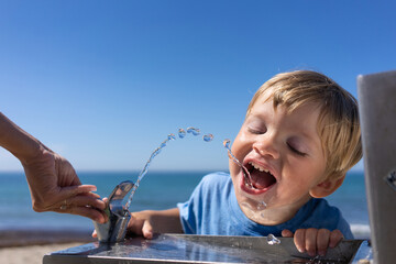 blond boy 3 years old drinks water from a water fountain on the beach in summer