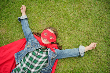 Girl Plays Superhero. Happy girl are playing superhero. Kid run across green field in red cloak at...