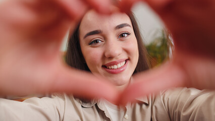 I love you. Happy young woman at home living room couch makes symbol of love, showing heart sign to camera, express romantic feelings express sincere positive feelings. Charity, gratitude, donation