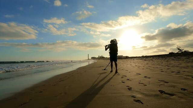Man with metal detector searching for precious objects and coins at beach and sea enjoying colorful sunset with cloudy sky
